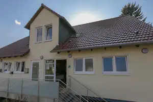 Ferienhaus am Campingplatz mit Terrasse, Geländer und Fenstern unter einem Satteldach, im Hintergrund Bäume.