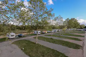Campingstellplätze mit Wohnwagen und Autos, gepflasterter Fahrweg und Rasenflächen zwischen Bäumen.