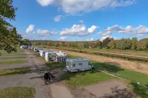 Wohnwagen auf einem Campingstellplatz entlang eines Graswegs, weitere Fahrzeuge in der Reihe, Bäume und Wiese im Hintergrund.