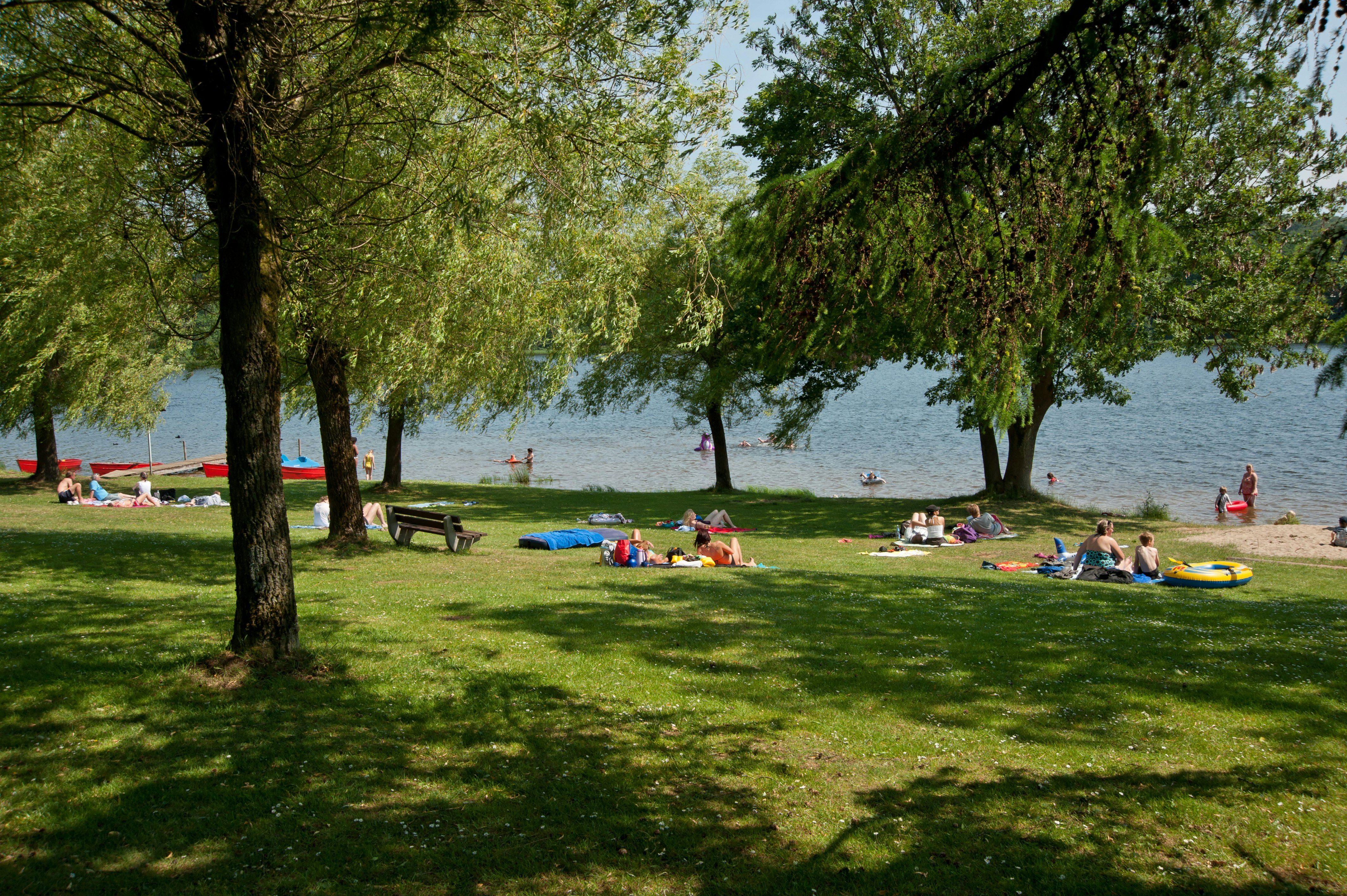 Grüner Campingplatz am See: Liegewiese mit Decken, Sonnenschirmen und Picknicktischen unter Bäumen.