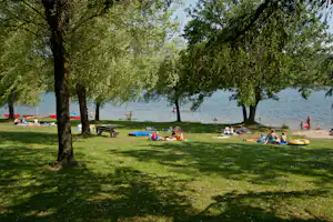 Grüner Campingplatz am See: Liegewiese mit Decken, Sonnenschirmen und Picknicktischen unter Bäumen.