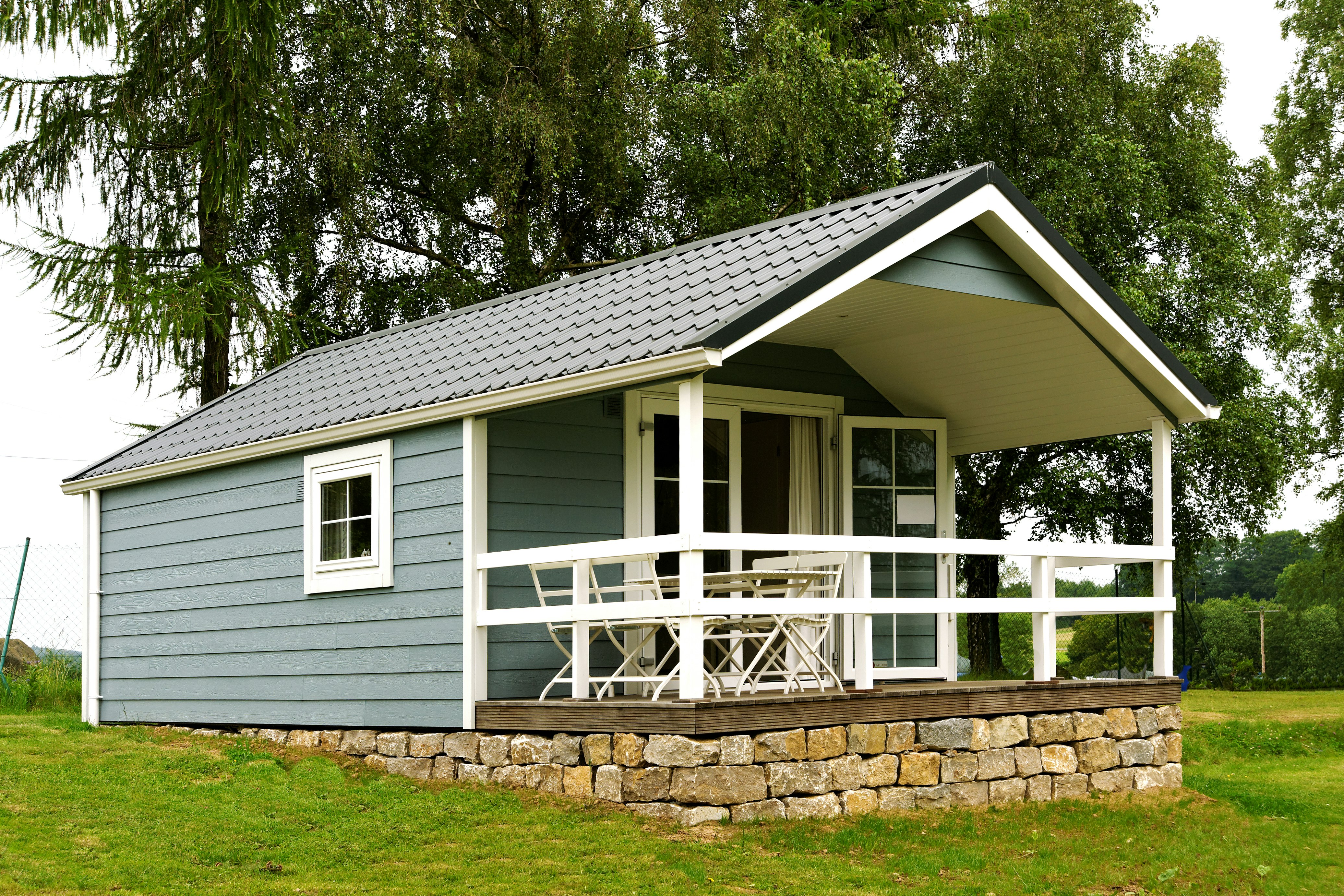 Modernes Campinghaus mit Holzveranda und Geländer, umgeben von Rasen und einem niedrigen Natursteinsockel, vor Bäumen.