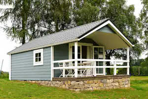 Modernes Campinghaus mit Holzveranda und Geländer, umgeben von Rasen und einem niedrigen Natursteinsockel, vor Bäumen.