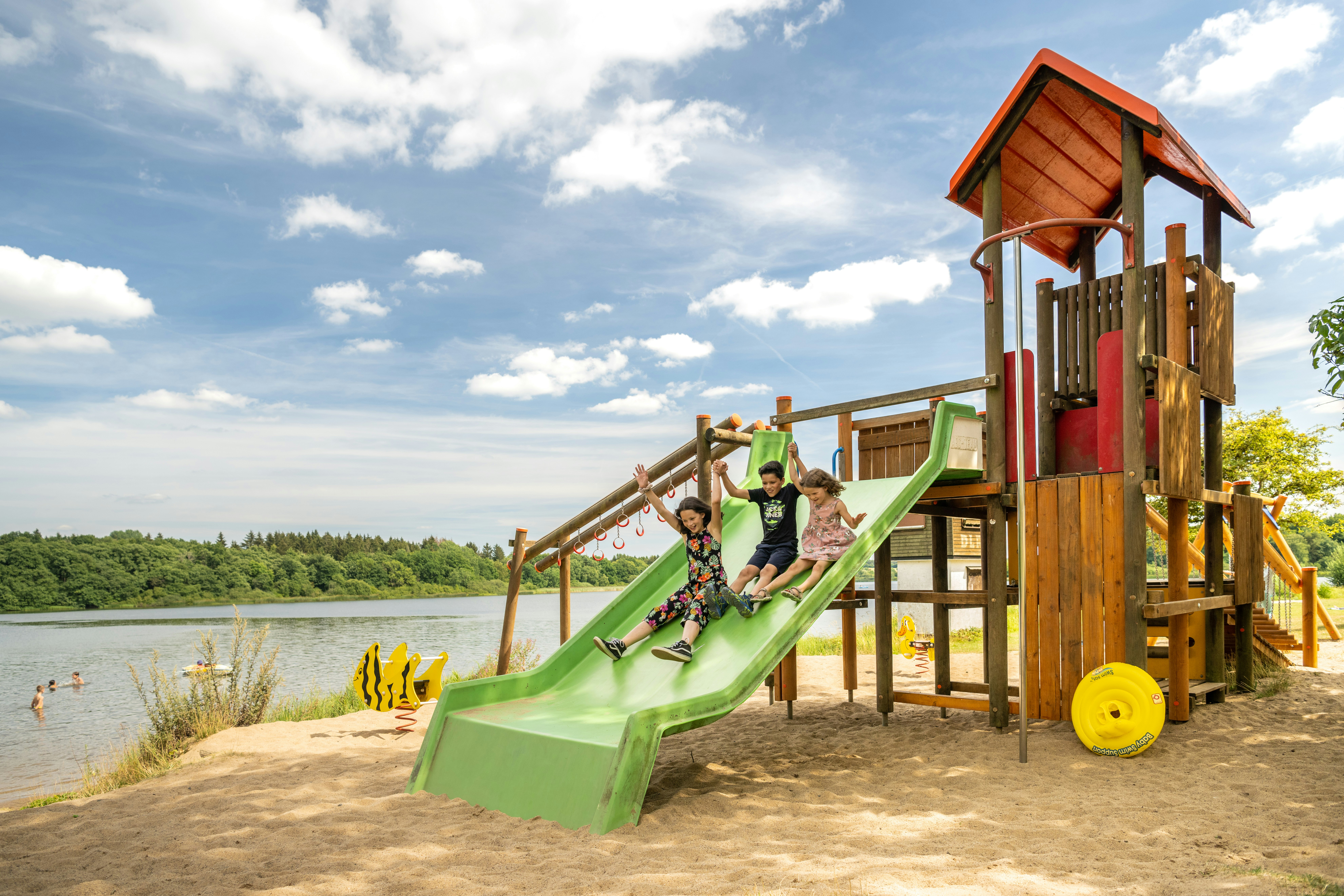 Spielplatz am See mit hölzernem Kletterturm und Rutsche, Sandboden und Blick auf Wasser und Bäume.