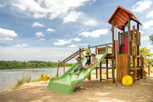Spielplatz am See mit hölzernem Kletterturm und Rutsche, Sandboden und Blick auf Wasser und Bäume.
