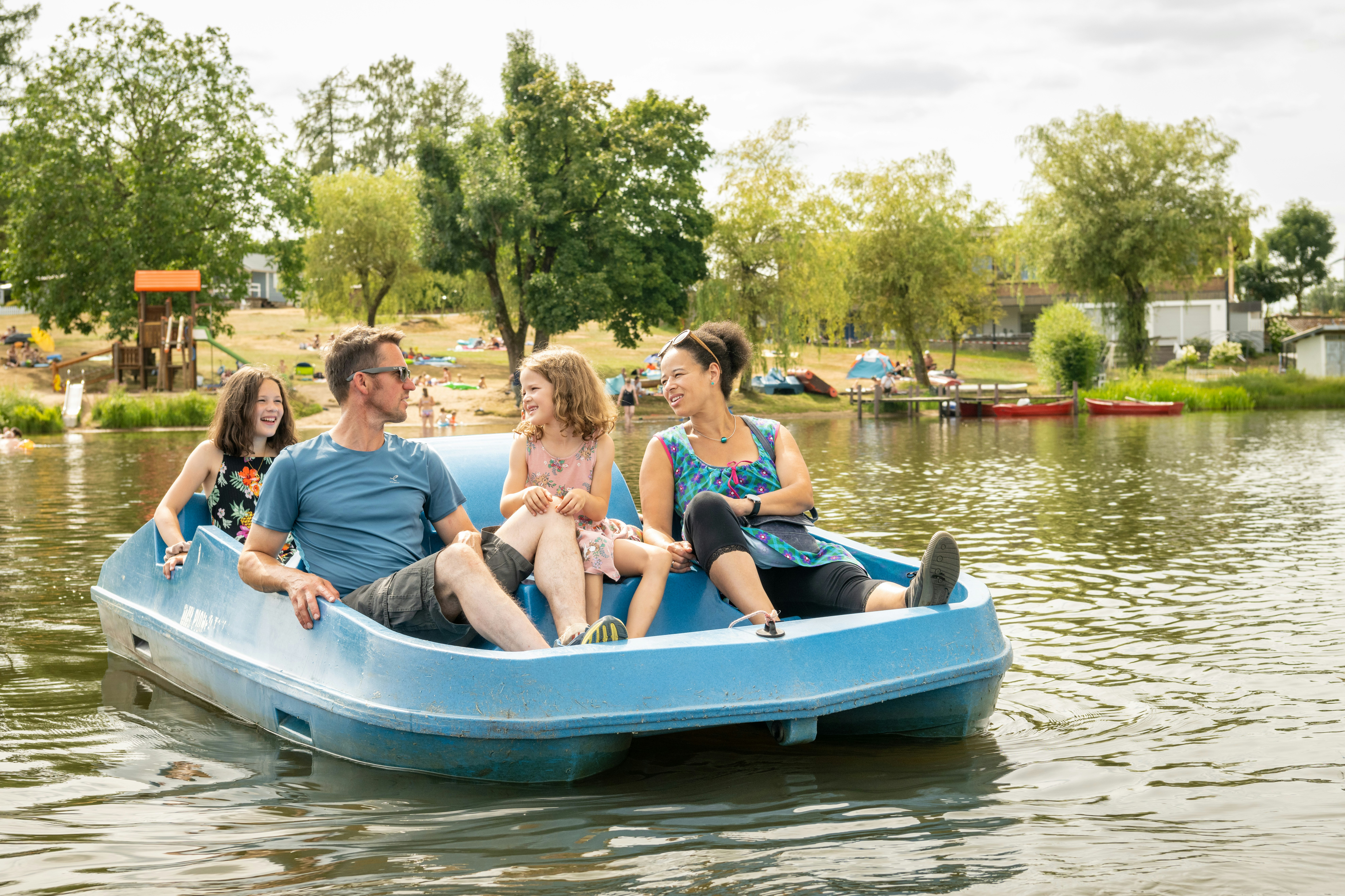 Blaues Ruderboot auf dem See mit vier Personen, im Hintergrund Bäume, Campingplatz und weitere Boote.