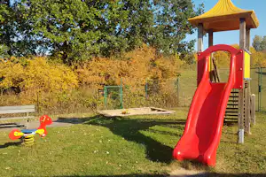 Rote Rutsche und Spielgerät auf einem Campingplatz-Spielplatz, mit Holzpodest, Zaun und Bäumen im Hintergrund.