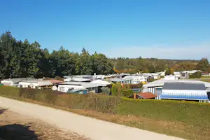 Überblick über einen Campingplatz mit Wohnwagen, Markisen und gepflegten Hecken am Weg, im Hintergrund Wald und Himmel.