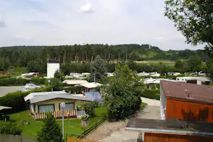 Campinganlage mit kleinen Hütten, gepflastertem Weg und Hecken, im Hintergrund Wald und grüne Hügel.