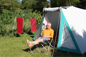 Campingzelt im Grünen mit Liegestuhl und Person im Vordergrund, Wäscheleine mit roten Tüchern im Hintergrund.
