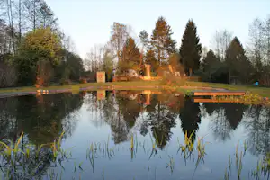 Teich mit Spiegelung, Holzsteg und Sitzbank am Campingplatz, umgeben von Bäumen und Rasenfläche.