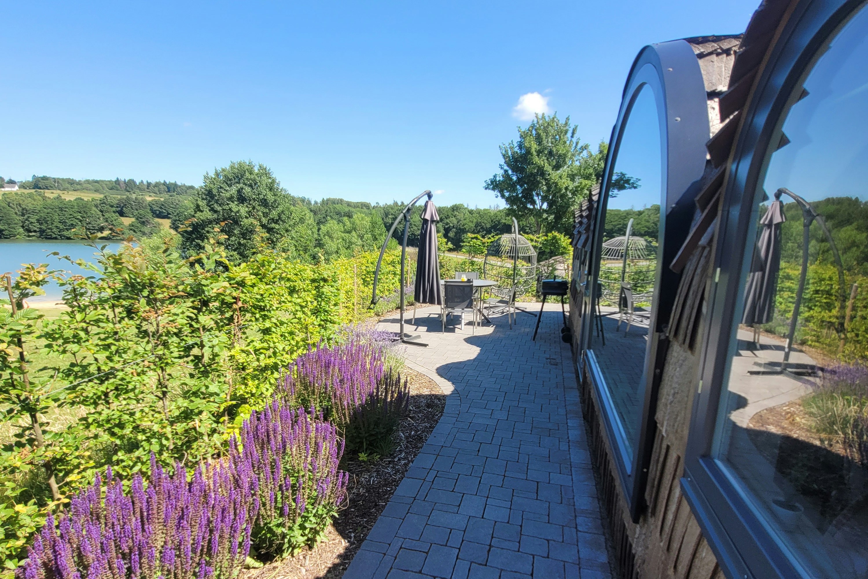 Stellplatz am Campingplatz mit gepflastertem Weg neben einem Wohnwagen, Lavendelbeeten und Blick auf den See in der Landschaft.