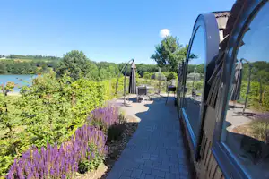 Stellplatz am Campingplatz mit gepflastertem Weg neben einem Wohnwagen, Lavendelbeeten und Blick auf den See in der Landschaft.