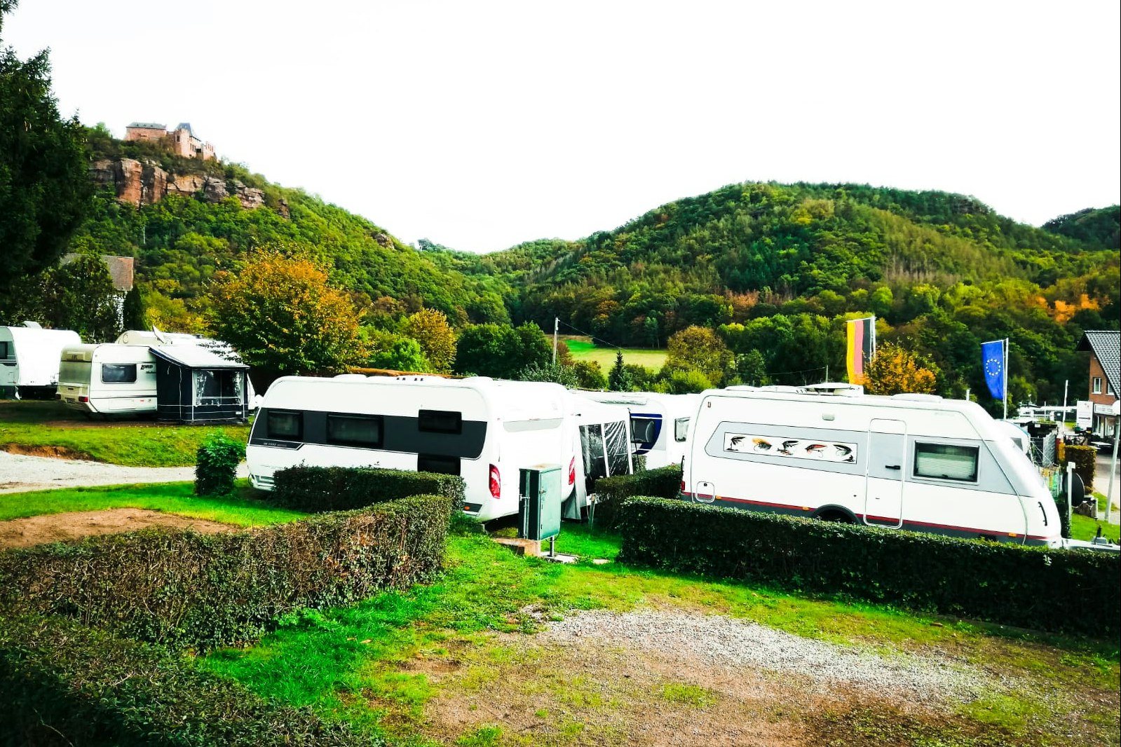 Mehrere Wohnmobile auf einem Campingplatz, umgeben von Hecken und Grünflächen, im Hintergrund bewaldete Hügel und ein Gebäude.