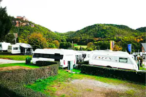 Mehrere Wohnmobile auf einem Campingplatz, umgeben von Hecken und Grünflächen, im Hintergrund bewaldete Hügel und ein Gebäude.