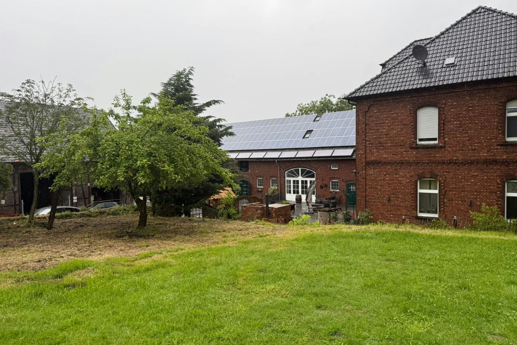 Backsteingebäude mit Solarpaneelen, Rasen und Bäumen im Garten, Terrasse mit Stühlen und Tisch vor den Fenstern.