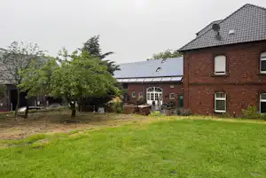Backsteingebäude mit Solarpaneelen, Rasen und Bäumen im Garten, Terrasse mit Stühlen und Tisch vor den Fenstern.