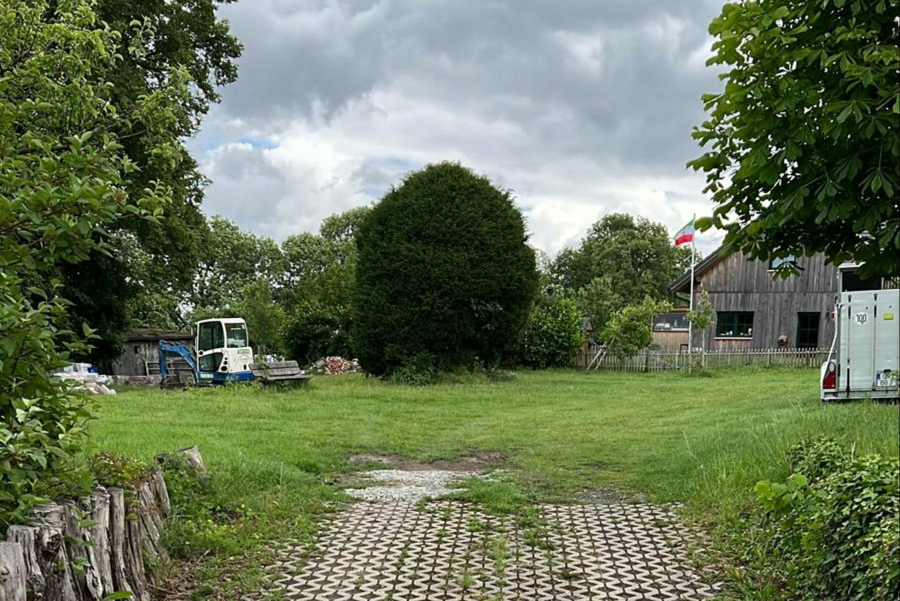 Grüner Campingplatz mit gepflastertem Weg, Rasenfläche, Bäumen und einem Gebäude am Rand.