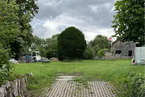 Grüner Campingplatz mit gepflastertem Weg, Rasenfläche, Bäumen und einem Gebäude am Rand.