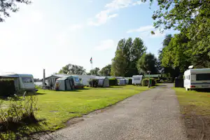 Reihe von Wohnwagen und Zelten auf einem Campingplatz, mit Rasenfläche, Kiesweg und Bäumen am Rand.