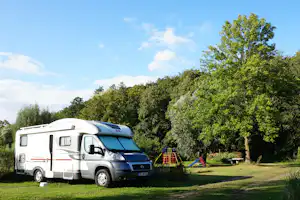 Wohnmobil auf einem Campingplatz im Grünen, mit Rasenfläche, Bäumen und Spielgeräten im Hintergrund.