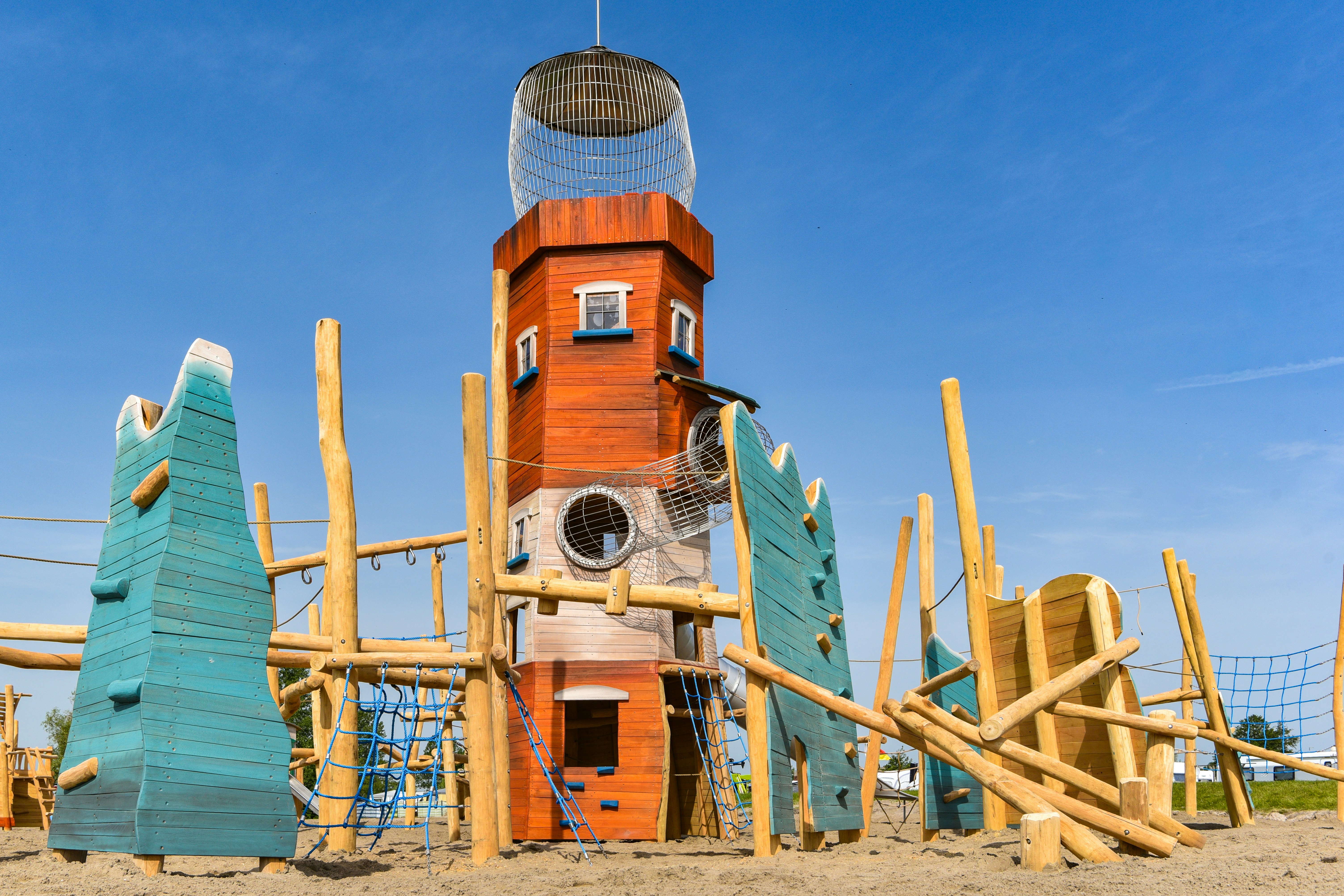 Holzspielturm mit Netz und Rutsche auf einem Campingplatz, mit bunten Holzpaneelen und Zaun vor blauem Himmel.