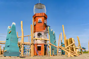 Holzspielturm mit Netz und Rutsche auf einem Campingplatz, mit bunten Holzpaneelen und Zaun vor blauem Himmel.