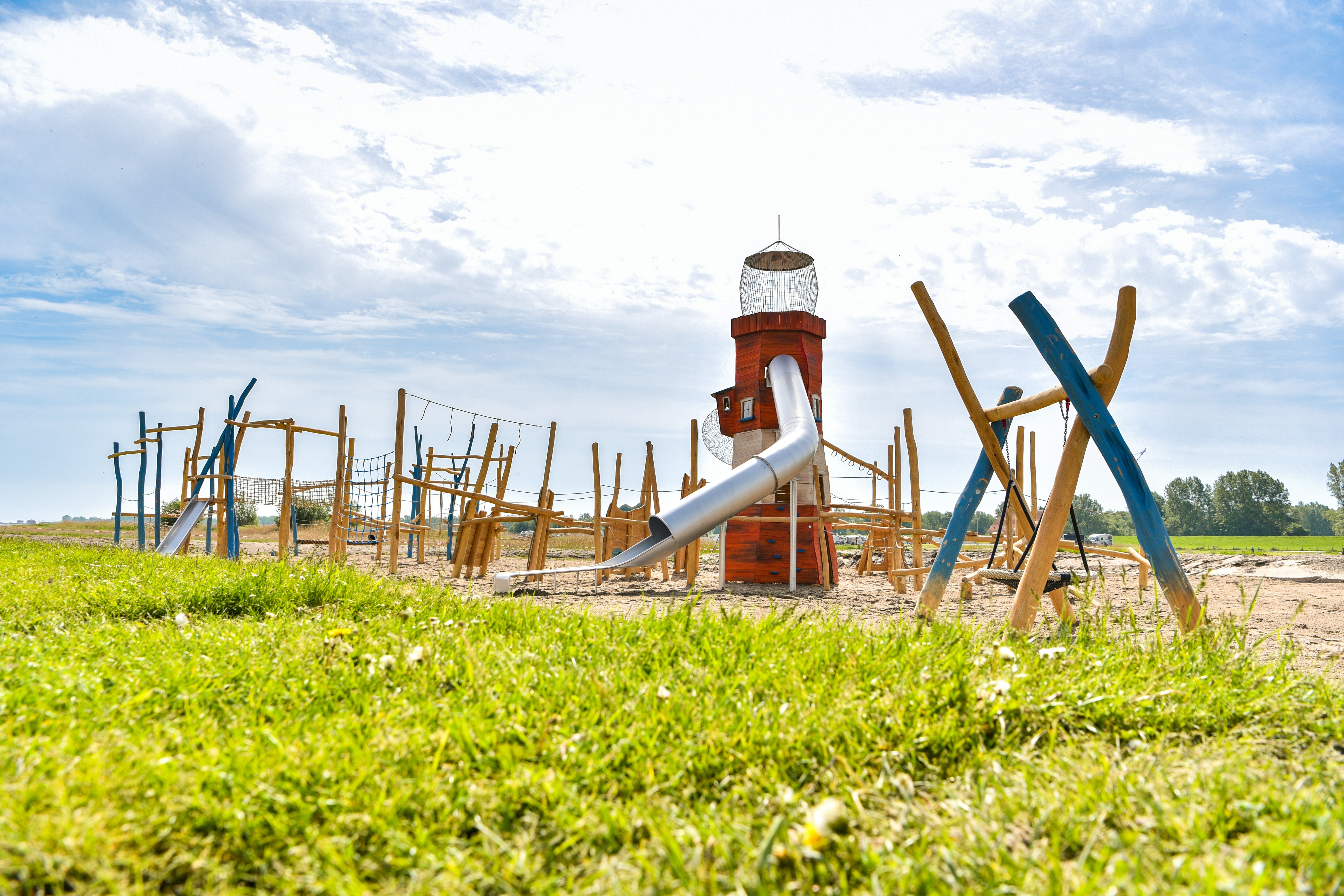 Ein Spielplatz mit Wasserspiel und Holzgeräten vor grünem Gras, im Hintergrund ein Himmel mit Wolken.