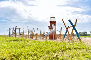 Ein Spielplatz mit Wasserspiel und Holzgeräten vor grünem Gras, im Hintergrund ein Himmel mit Wolken.