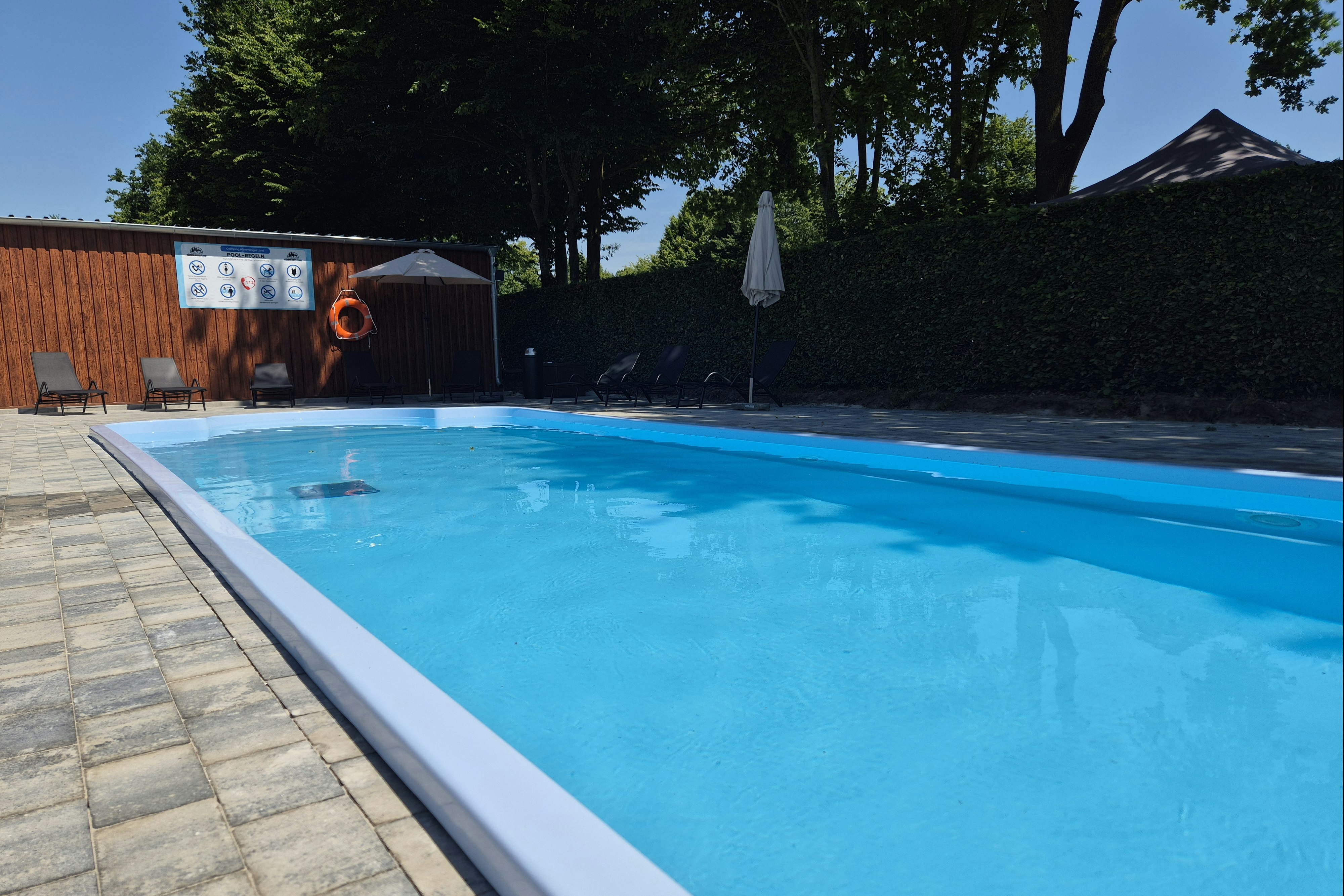 Schwimmbecken mit blauem Wasser und weißen Kanten, daneben gepflasterter Weg und Bäume vor einem Campingplatz-Gebäude.