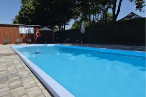 Schwimmbecken mit blauem Wasser und weißen Kanten, daneben gepflasterter Weg und Bäume vor einem Campingplatz-Gebäude.