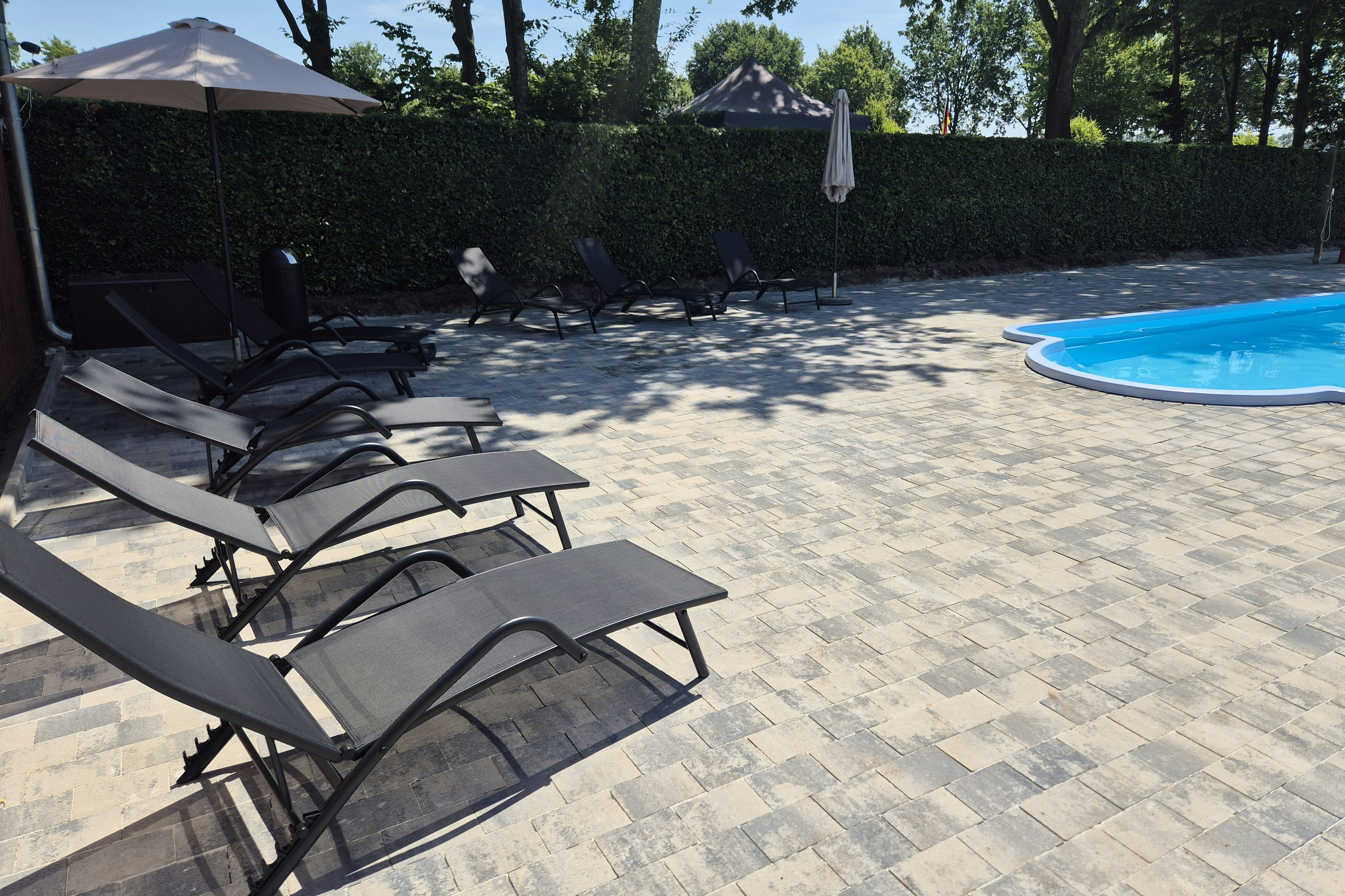 Sonnenliegen am gepflasterten Bereich neben einem Swimmingpool, umgeben von Hecke und Bäumen, im Hintergrund Gebäude und Zelt.