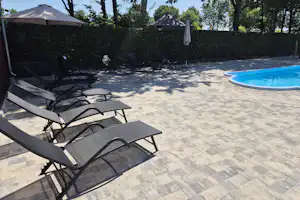 Sonnenliegen am gepflasterten Bereich neben einem Swimmingpool, umgeben von Hecke und Bäumen, im Hintergrund Gebäude und Zelt.
