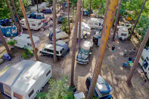 Campingplatz zwischen Bäumen: Wohnmobile, Zelte und Autos auf einem Kiesweg, mit Campingstühlen und Tischen.