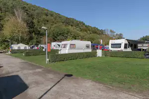 Wohnwagen auf einem Campingplatz, mit Rasenfläche, Hecken und gepflastertem Weg; im Hintergrund Bäume und Hügel.