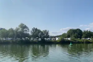 Seeufer mit Wohnmobilen und Mobilheimen am Campingplatz, dahinter Bäume und ein grüner Steg im Wasser.