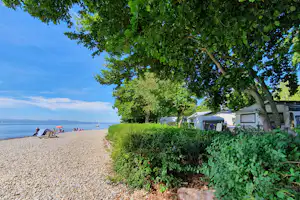 Campingplatz am See: Kiesstrand, Zelte und Wohnwagen unter großen Bäumen, im Hintergrund Boote auf dem Wasser.