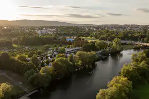 Panoramablick auf einen Fluss mit Uferwegen, Bäumen und einem Campingbereich in einem Tal mit Häusern in der Ferne.