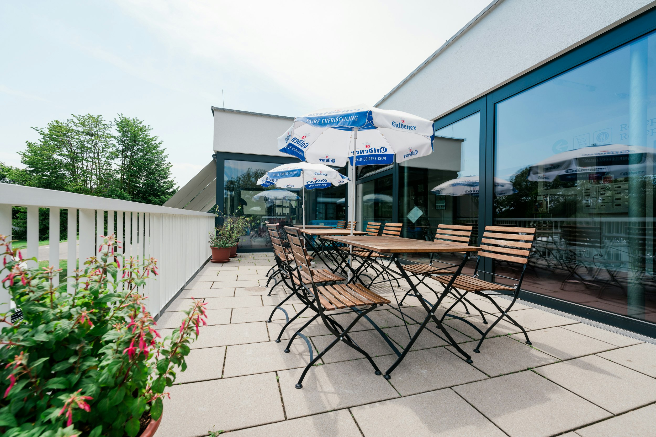 Terrasse am Campingplatz mit Tisch und Stühlen vor einem modernen Gebäude, Glasfront und Sonnenschirm, Hecken und Blumenbeete.