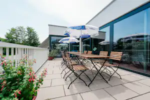 Terrasse am Campingplatz mit Tisch und Stühlen vor einem modernen Gebäude, Glasfront und Sonnenschirm, Hecken und Blumenbeete.