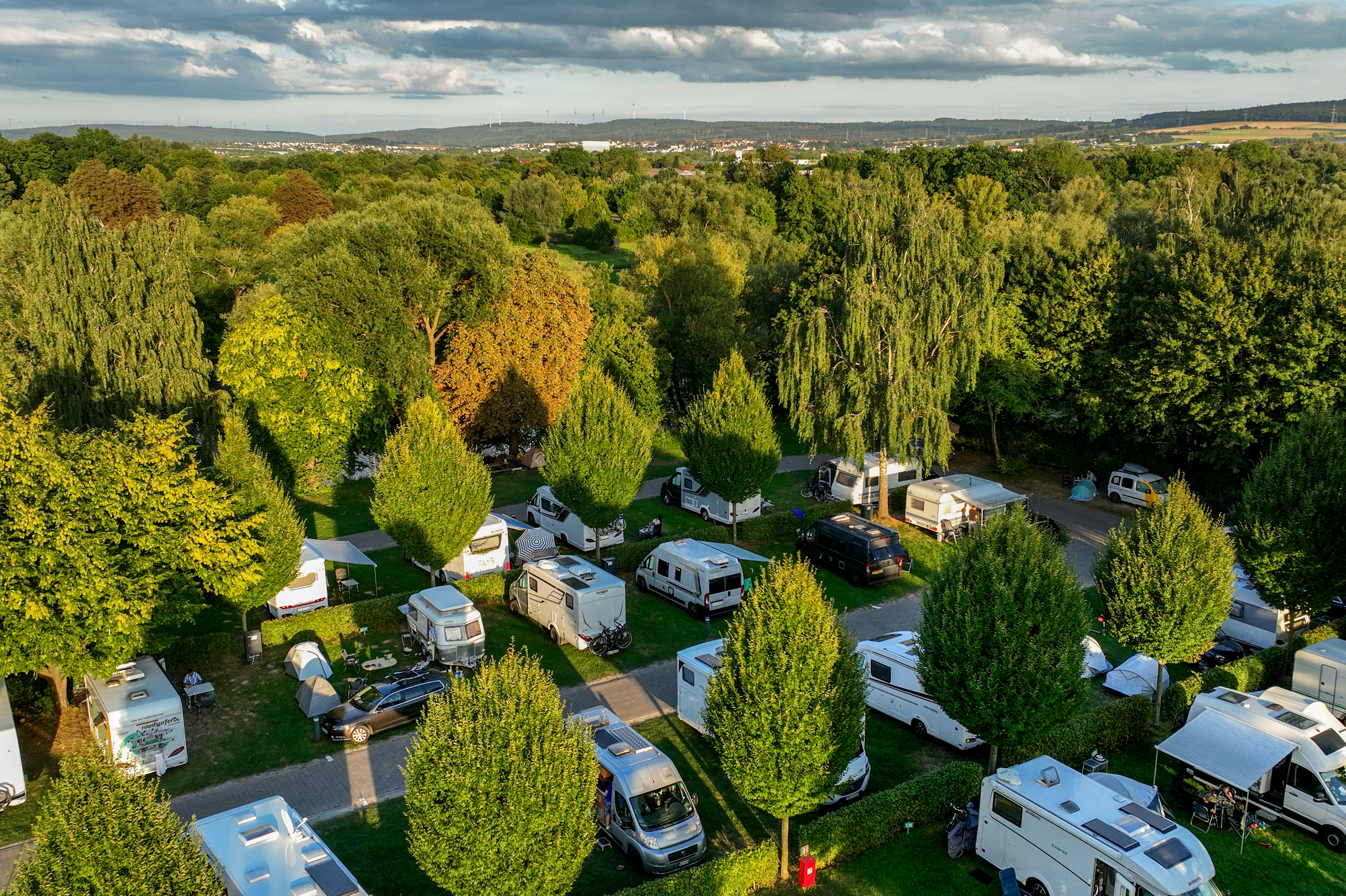Luftaufnahme eines Campingplatzes mit Stellplätzen, Wohnmobilen und Wohnwagen, Wegen und Bäumen in einer grünen Umgebung.