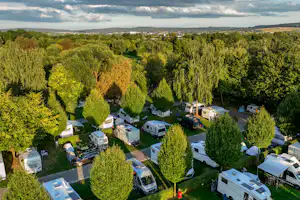 Luftaufnahme eines Campingplatzes mit Stellplätzen, Wohnmobilen und Wohnwagen, Wegen und Bäumen in einer grünen Umgebung.