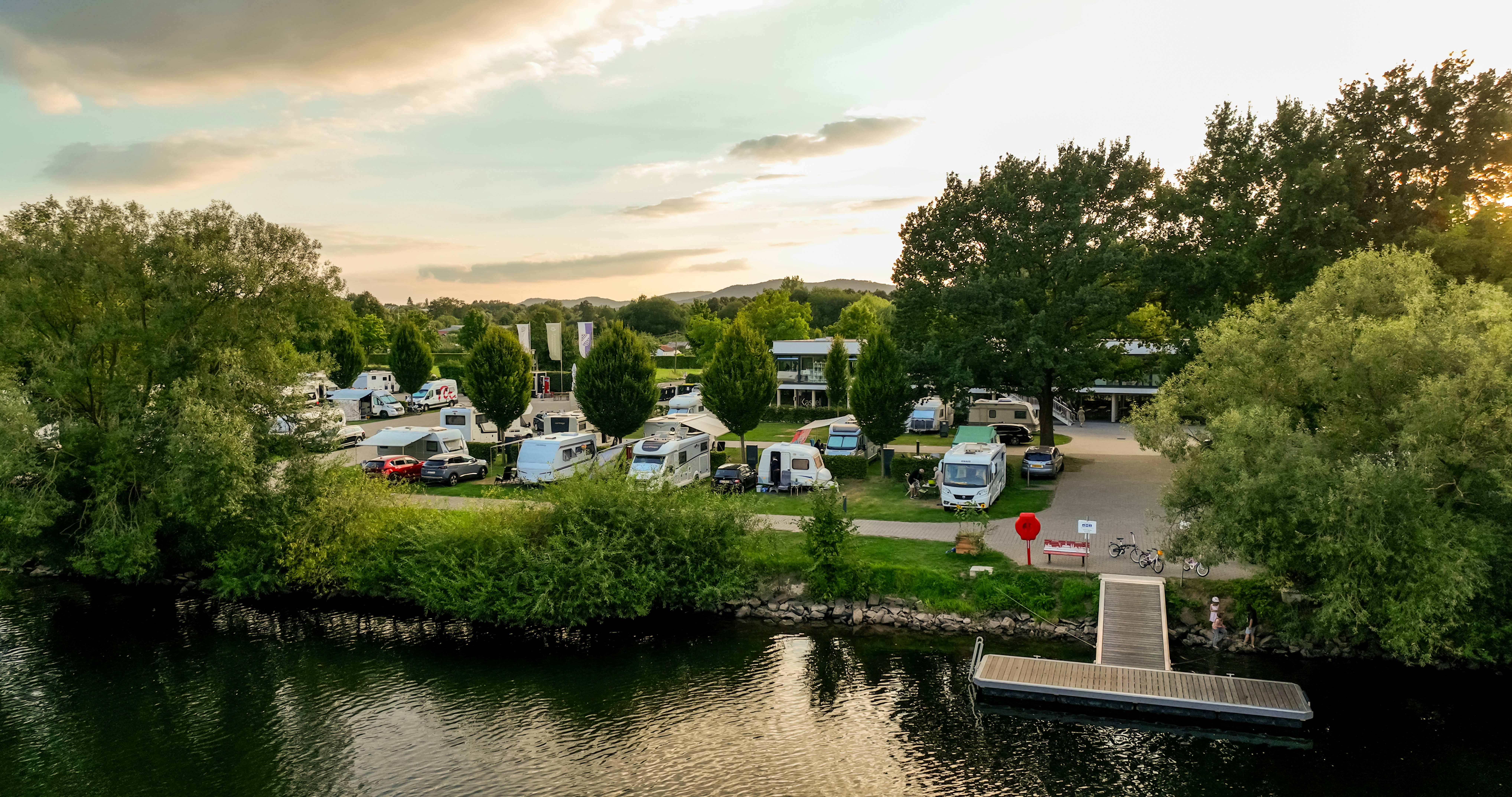 Campingplatz am Fluss mit Bootssteg, Stellplätzen und Wohnmobilen, umgeben von Bäumen und gepflegten Wegen.
