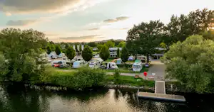 Campingplatz am Fluss mit Bootssteg, Stellplätzen und Wohnmobilen, umgeben von Bäumen und gepflegten Wegen.