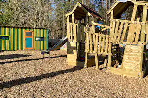 Holzspielgeräte mit Kletterwand und Podest auf einem Kiesplatz, im Hintergrund bunte Spielwand und Bäume im Campingbereich.
