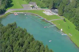 Blick auf einen See mit Schwimmstegen, Ruderbooten und Liegewiese, umgeben von Bäumen und einem Campingplatz mit Gebäuden.