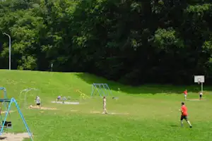 Spielplatz auf dem Campinggelände: Rasenfläche mit Schaukel, Sandbereich und Basketballkorb im Hintergrund.