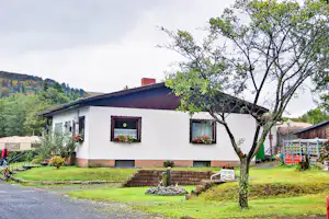 Weiße Ferienhaus-Fassade mit rotem Dach, Sitzbank und kleinem Garten; daneben Bäume, Weg und ein Fahrrad im Campingbereich.