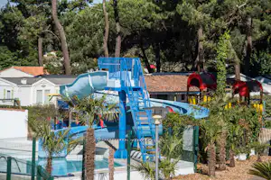 Blaue Wasserrutschen auf einem Campingplatz, umgeben von Palmen, Zaun und Bäumen, im Hintergrund Gebäude mit roten Dächern.