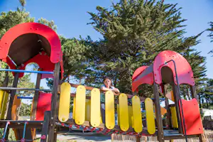 Roter und gelber Kletterturm mit Rutsche am Spielplatz, im Hintergrund Bäume und ein Kind auf der Plattform.